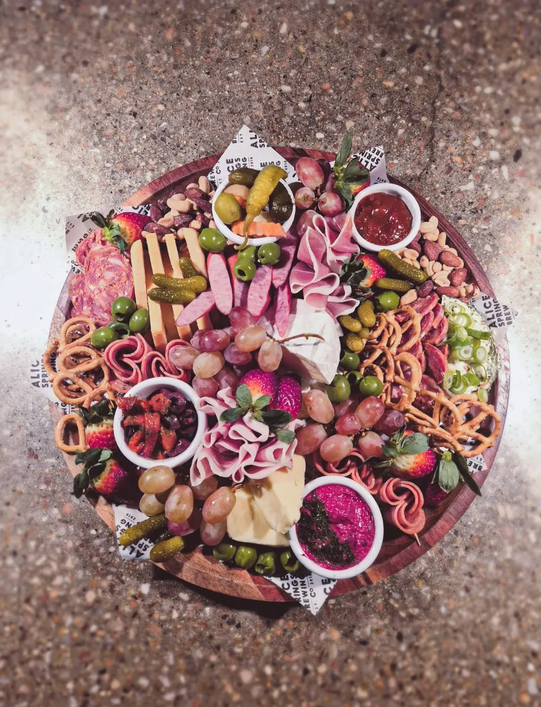 Overhead view of colourful antipasto grazing board with meats, olives, cheeses, dips and fruit on branded paper